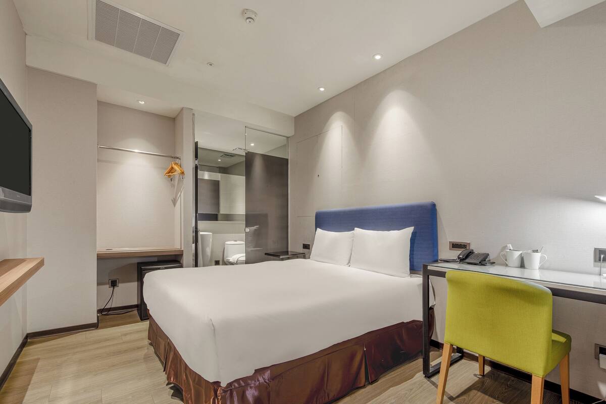 A hotel room with a bed, desk, chair, and a bathroom visible through a glass partition.