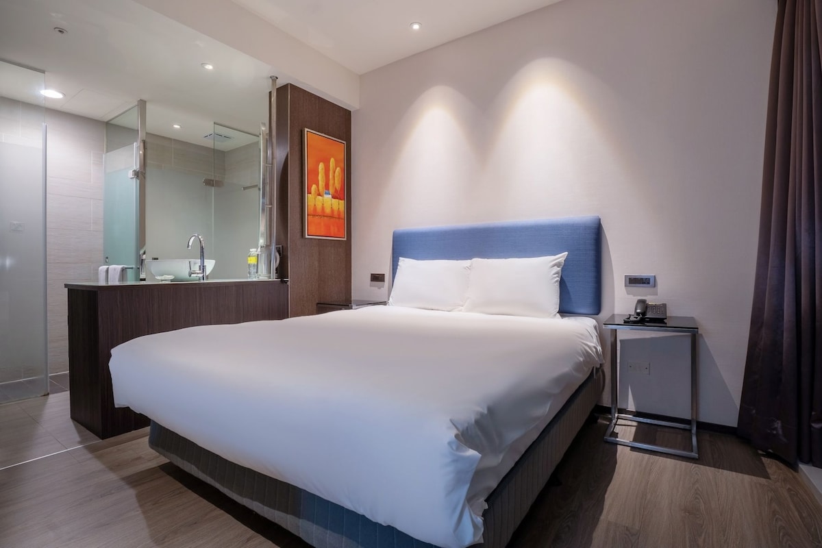 A modern hotel room with a large bed, a bedside table with a phone, a mirror, and a painting on the wall.