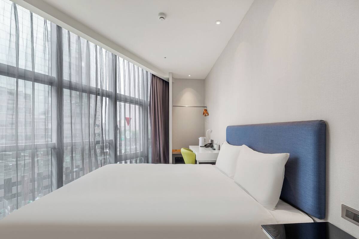 A modern hotel room with a large bed, a desk, and a view through sheer curtains.