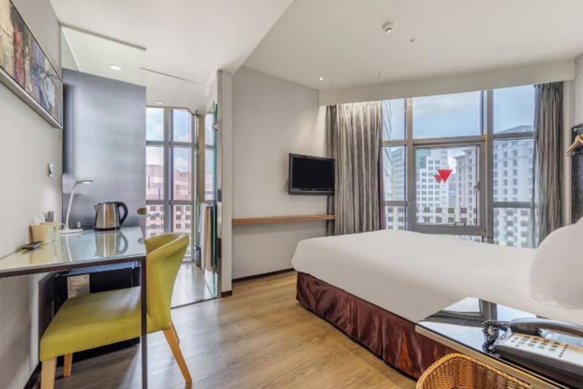 Habitación de hotel con cama, escritorio, silla, televisor y ventanas amplias.
