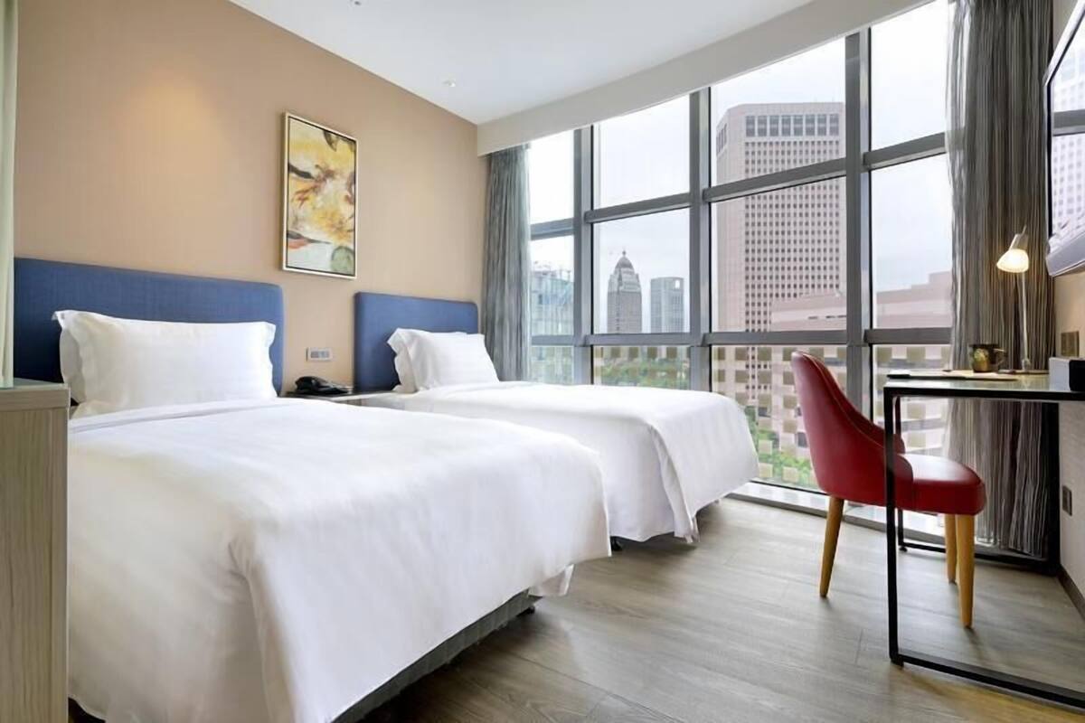 A hotel room with two beds, a desk with a chair, a large window with a city view, and a painting on the wall.