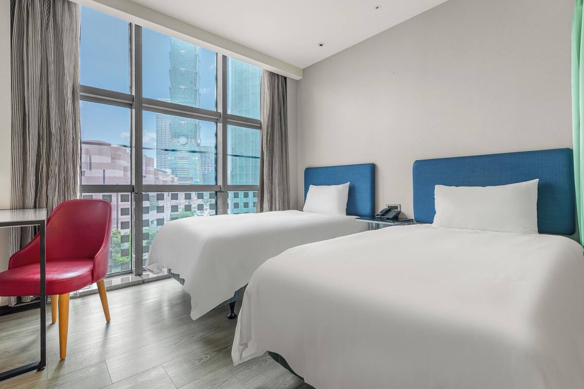 A hotel room with two beds, a red chair, a desk, and a large window with a city view.