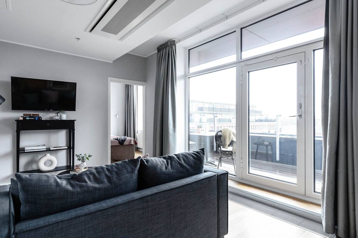 Een moderne woonkamer met een grijze bank, een flatscreen televisie en een groot raam met uitzicht op de stad.