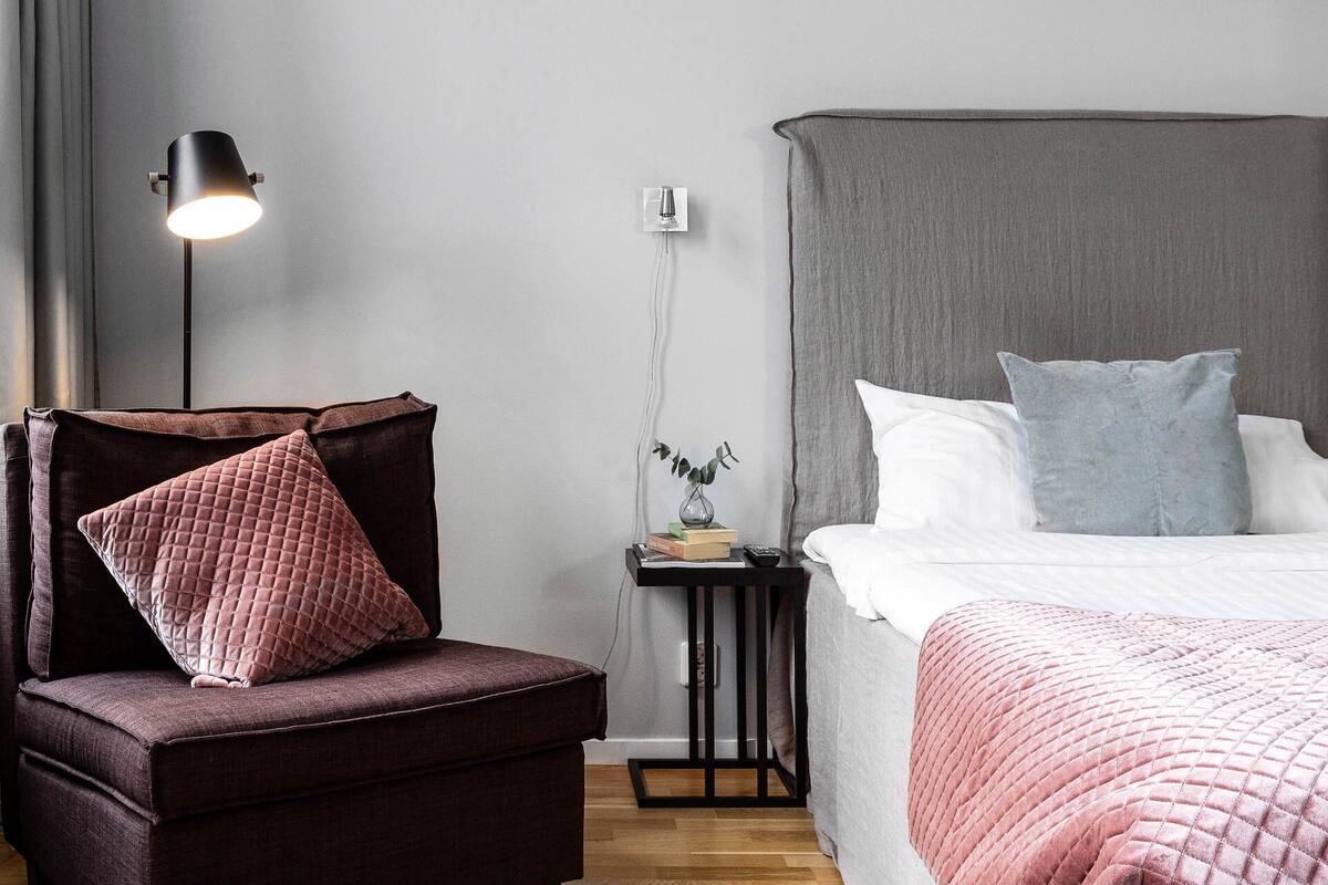 Een moderne slaapkamer met een donkerbruine fauteuil, een nachtkastje met een lamp, een kamerplant en een groot bed met een grijze hoofdbord.