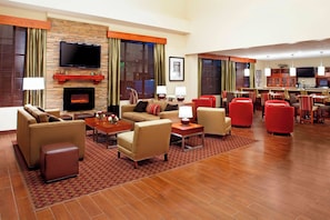 Un vestíbulo de hotel con chimenea, sofás, televisor y zona de comedor con sillas rojas.