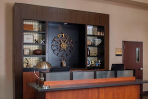 Un mostrador de recepción con un detalle metálico decorativo en la parte superior, un cuadro enmarcado y una lámpara a la izquierda.