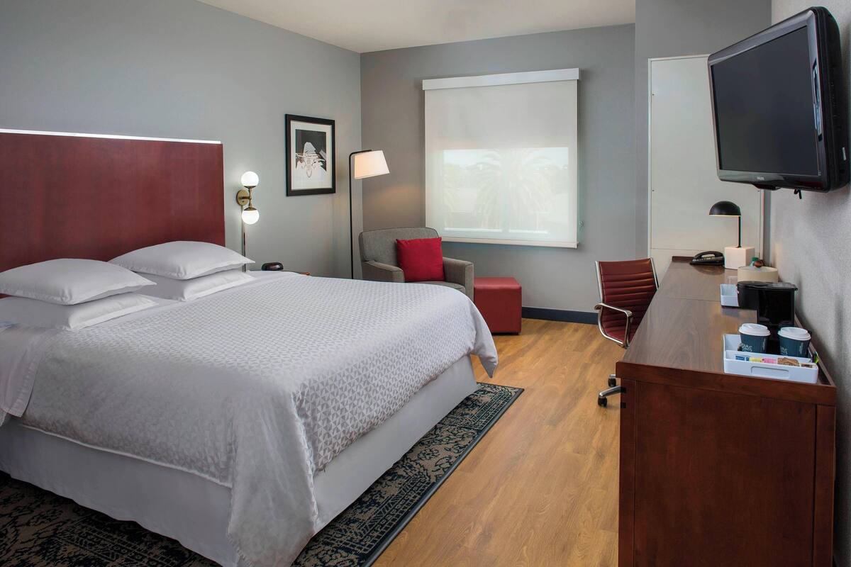A hotel room with a large bed, a desk with a computer, a TV, and a window with a view of palm trees.