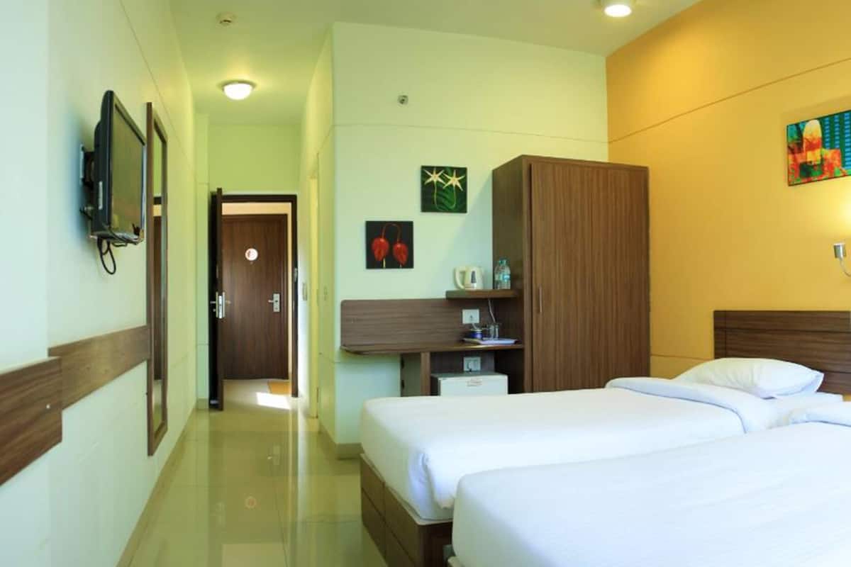 A hotel room with two beds, a wooden wardrobe, a desk with a chair, a small table with a kettle, and a wall-mounted TV.