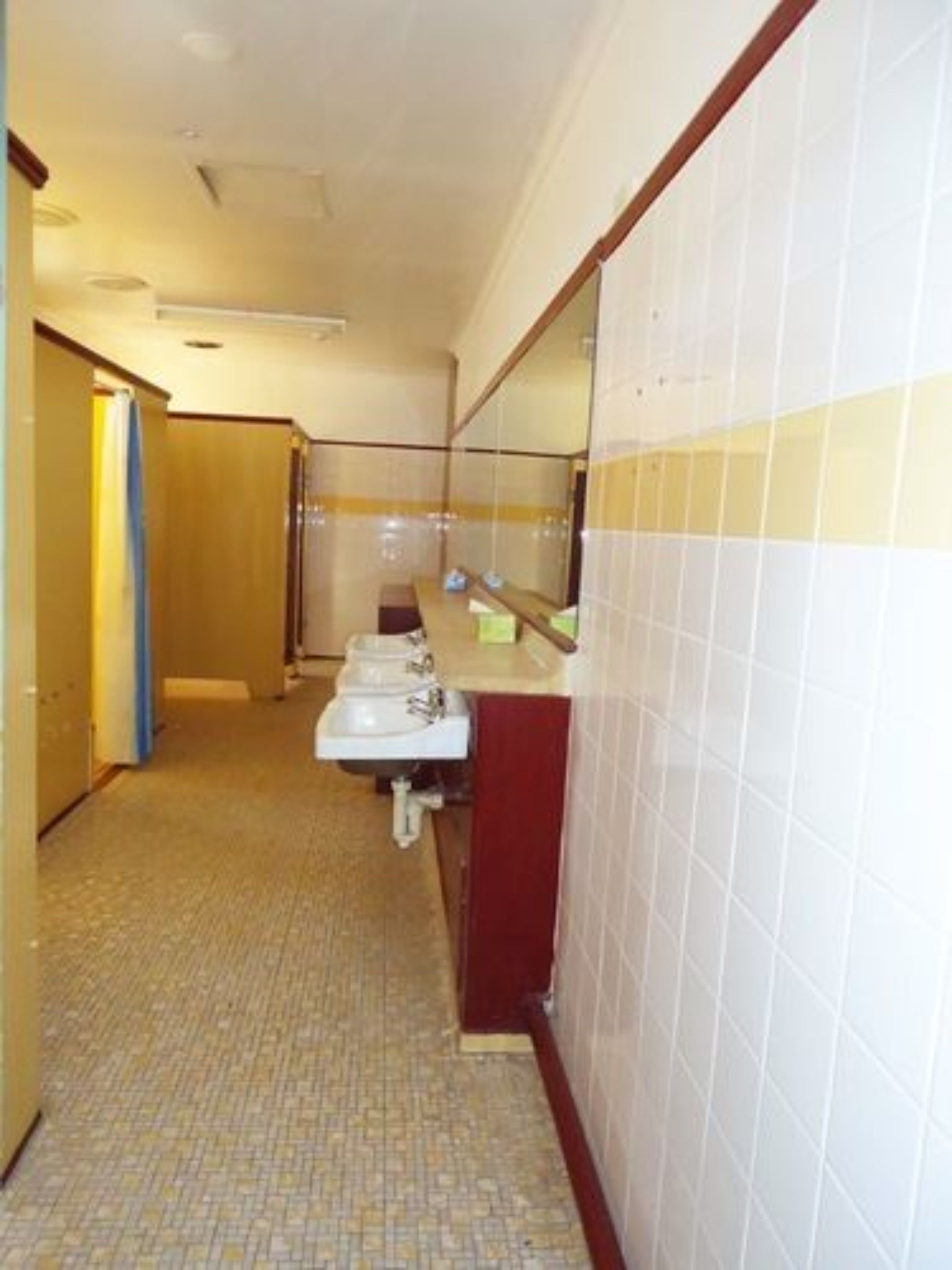 Un couloir étroit, dont le sol est recouvert de carreaux, mène à une salle de bain équipée de deux lavabos et d’un miroir.
