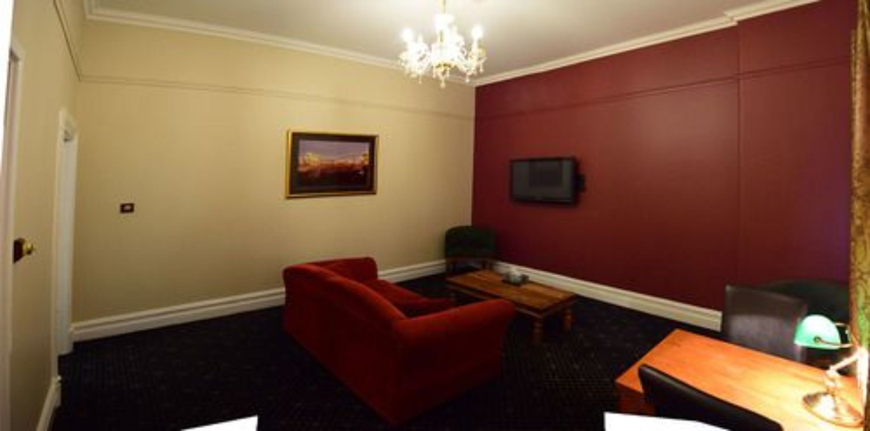 Ein Zimmer mit einem roten Sofa, einem kleinen Tisch, einem an der Wand befestigten Fernseher und einer Chandelüster.