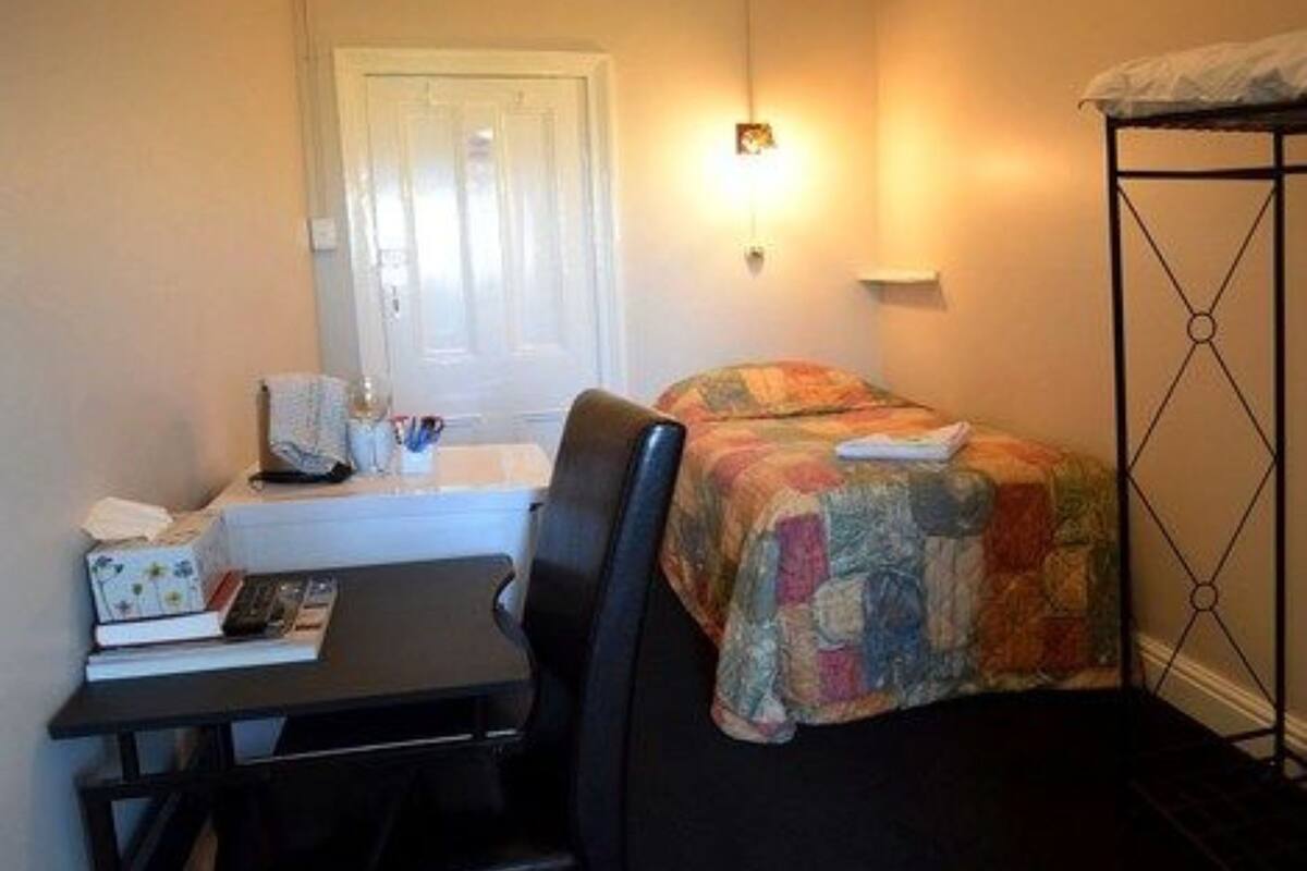 Une petite pièce, de taille modeste, comprenant un lit, un bureau et une chaise. On y trouve une fenêtre avec des rideaux, un luminaire fixé au mur et un radiateur.