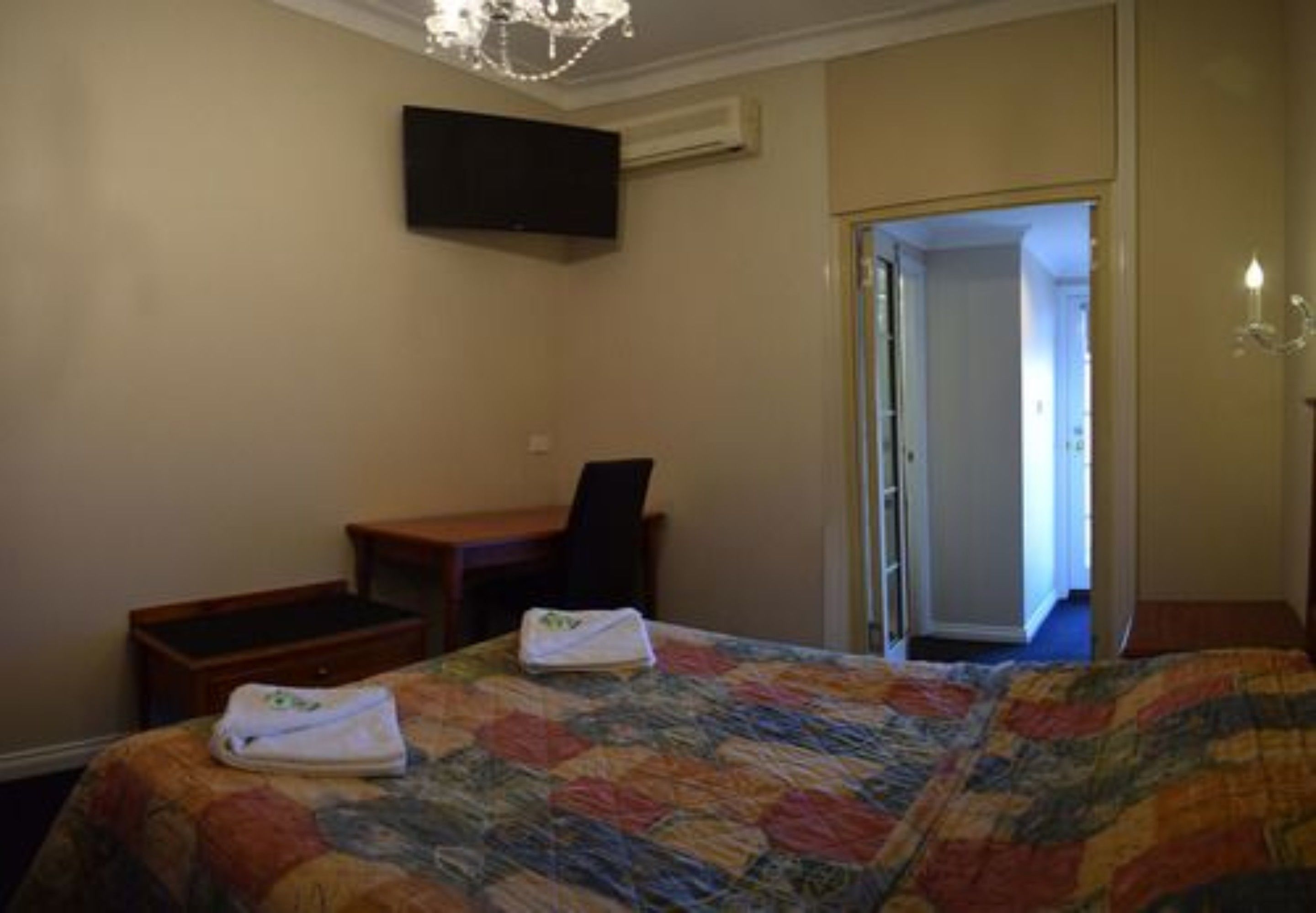 A hotel room with a bed, a desk, a chair, a TV mounted on the wall, and a door leading to another room.