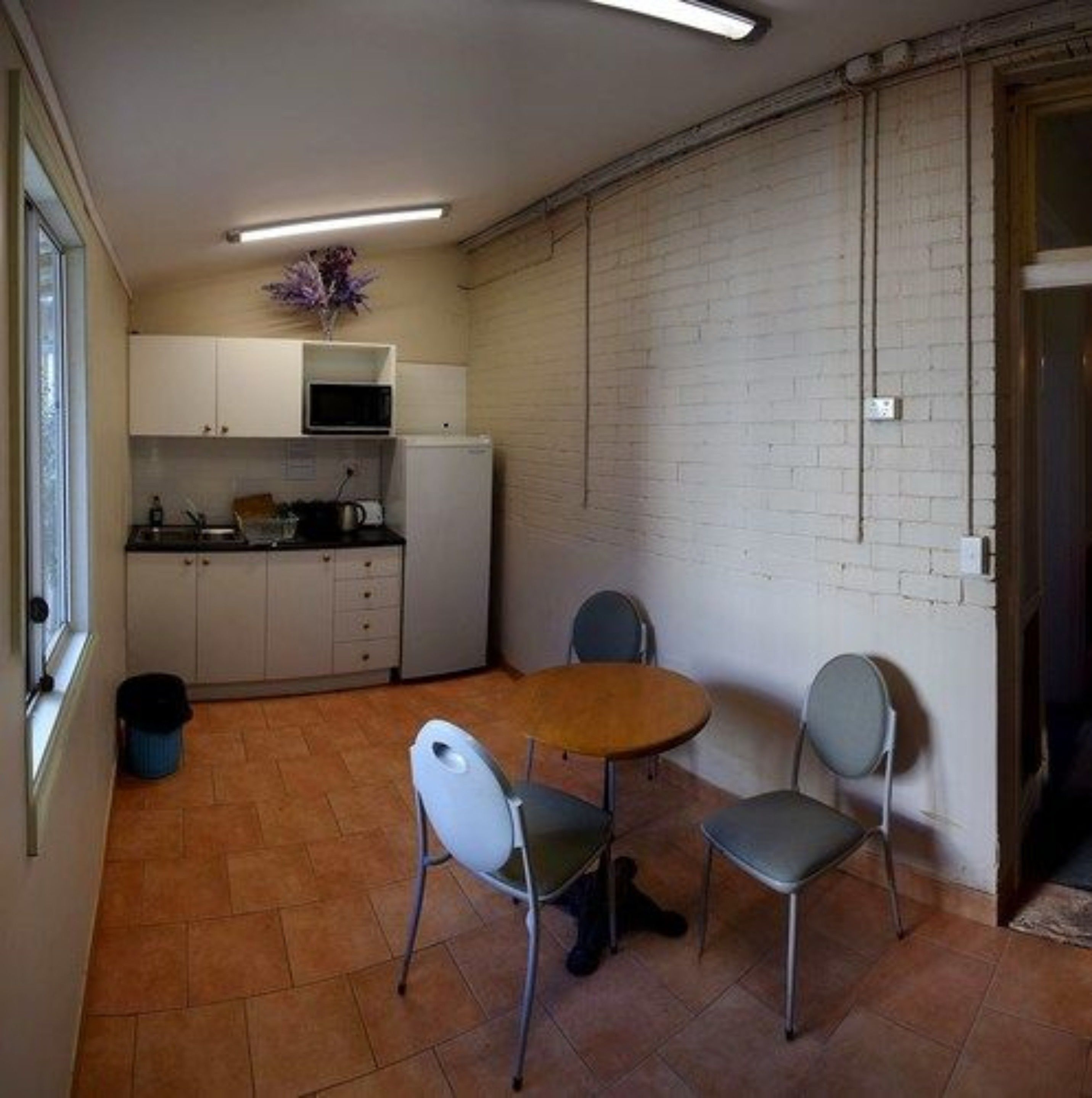 A compact kitchen with white cabinets, a microwave, and a small refrigerator. There is a dining table with two chairs and a potted plant on the counter.