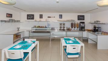A modern kitchen with white countertops, stainless steel appliances, and a coffee station. There are two tables with chairs, each set with a white cup, saucer, and utensils. The walls are decorated with motivational quotes and a beach-themed sign.