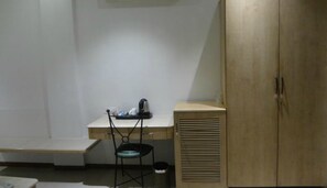 A small, dimly lit room with a single chair, a small table with a kettle, and a wooden wardrobe. - Hotel Suncity Apollo (Mumbai)