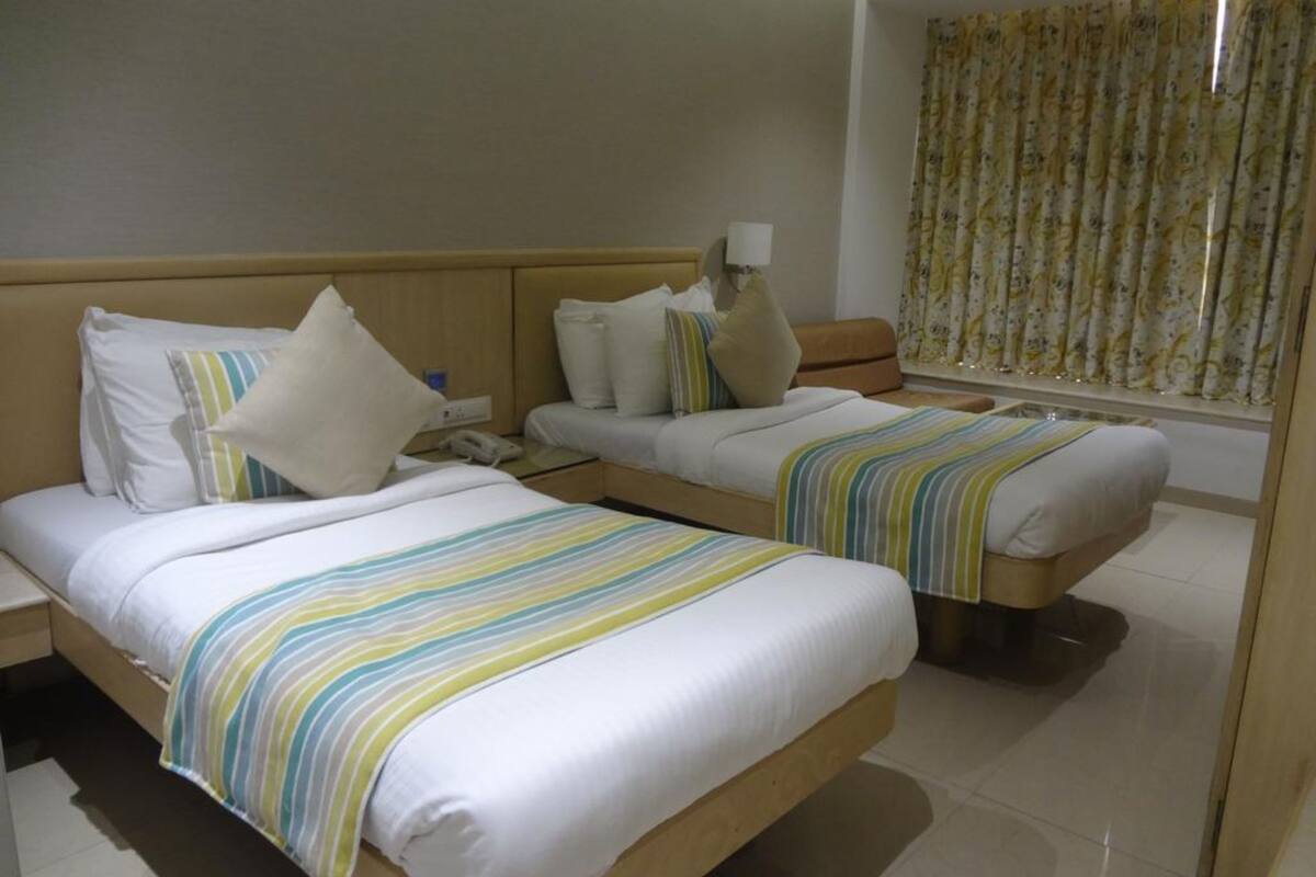 A hotel room with two beds, each with striped bedding and pillows. There is a bedside table with a lamp and a phone. The room has a window with floral curtains.