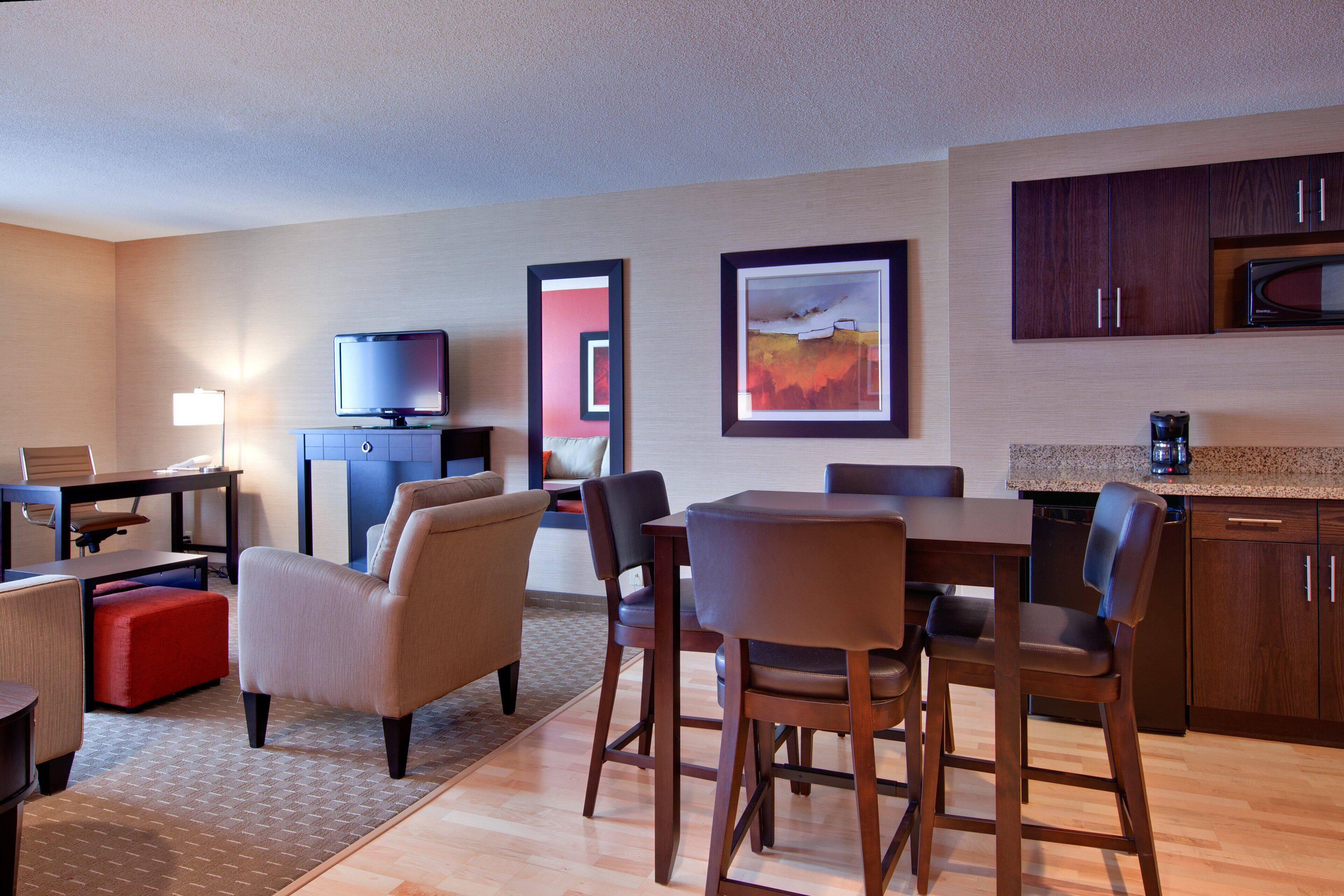 A hotel room with a dining area, a television, artwork on the wall, and wooden cabinets.