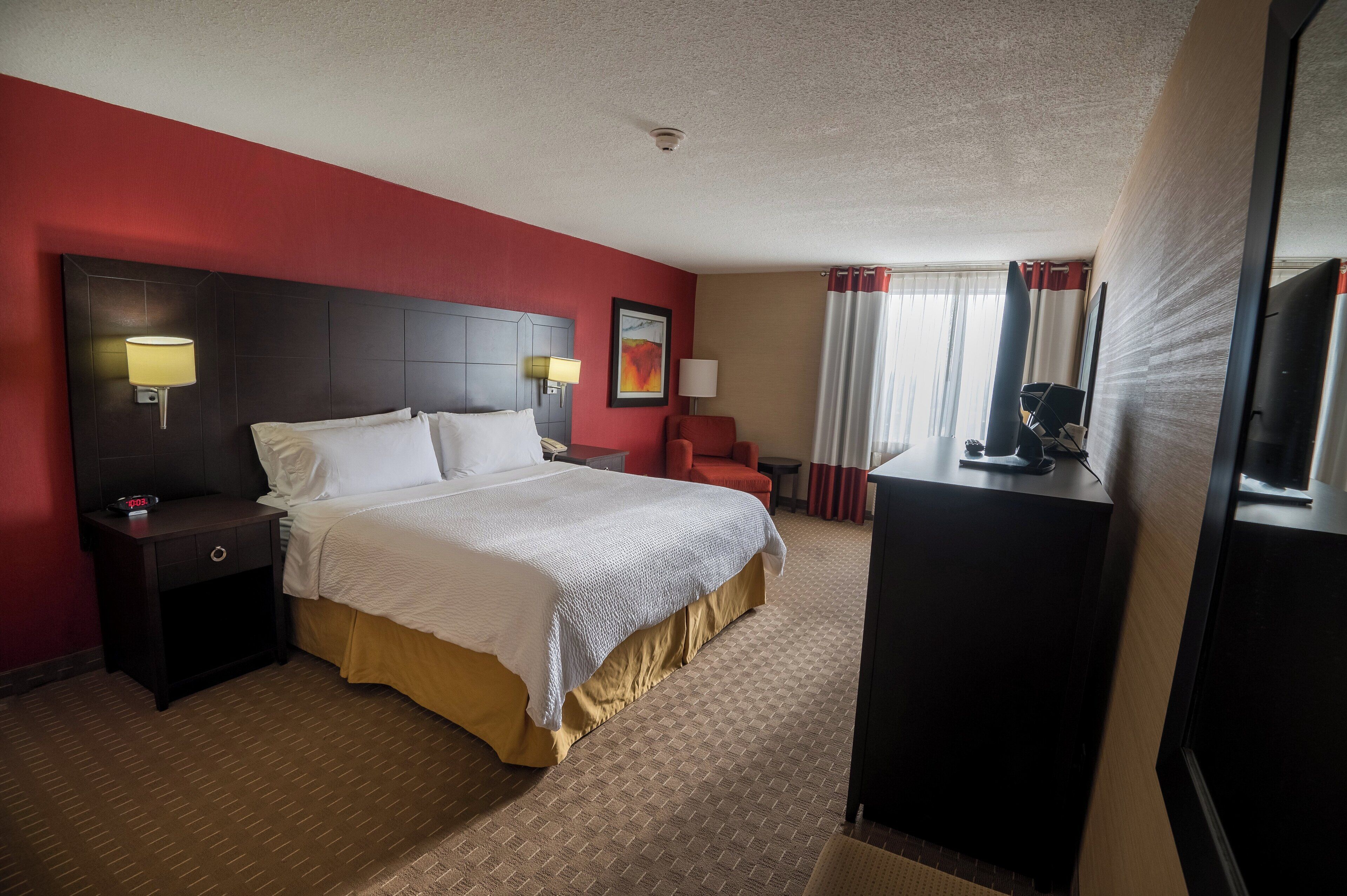 Quarto de hotel com uma cama grande, uma mesa de cabeceira com abajur, uma escrivaninha com cadeira, uma televisão e uma janela com cortinas.