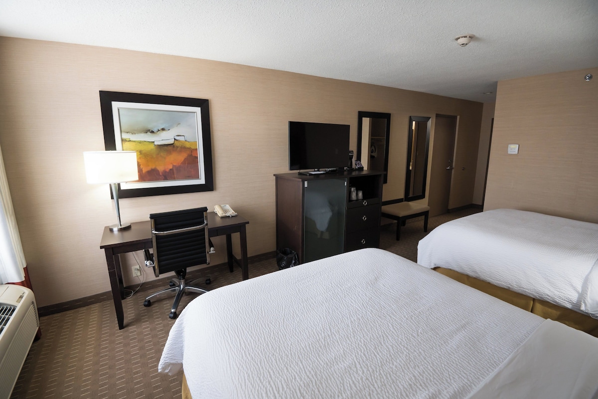 A hotel room with two beds, a desk with a telephone, a chair, a desk lamp, a flat-screen TV, a mirror, and a wall-mounted heater.
