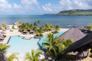 Zona de piscina en un resort con palmeras, estructuras de techo de paja y vista al mar.
