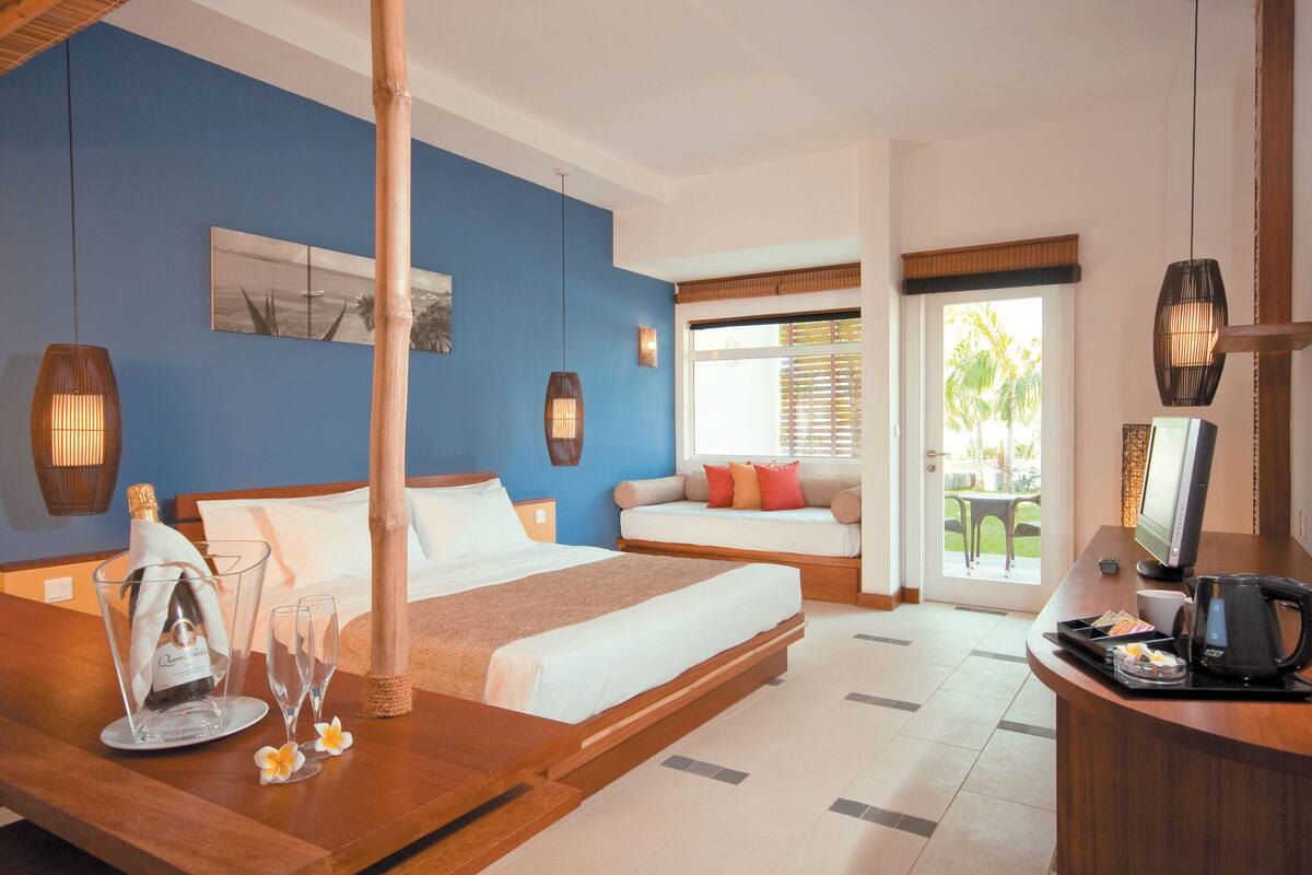 A hotel room with a bed, bedside table, and a desk with a computer. There is a window with blinds and a view of the outdoors.