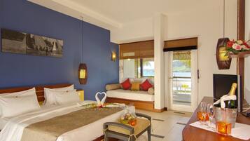 Habitación de hotel con cama, sofá, mesa de comedor con vasos y una botella, y vista al mar a través de la puerta.