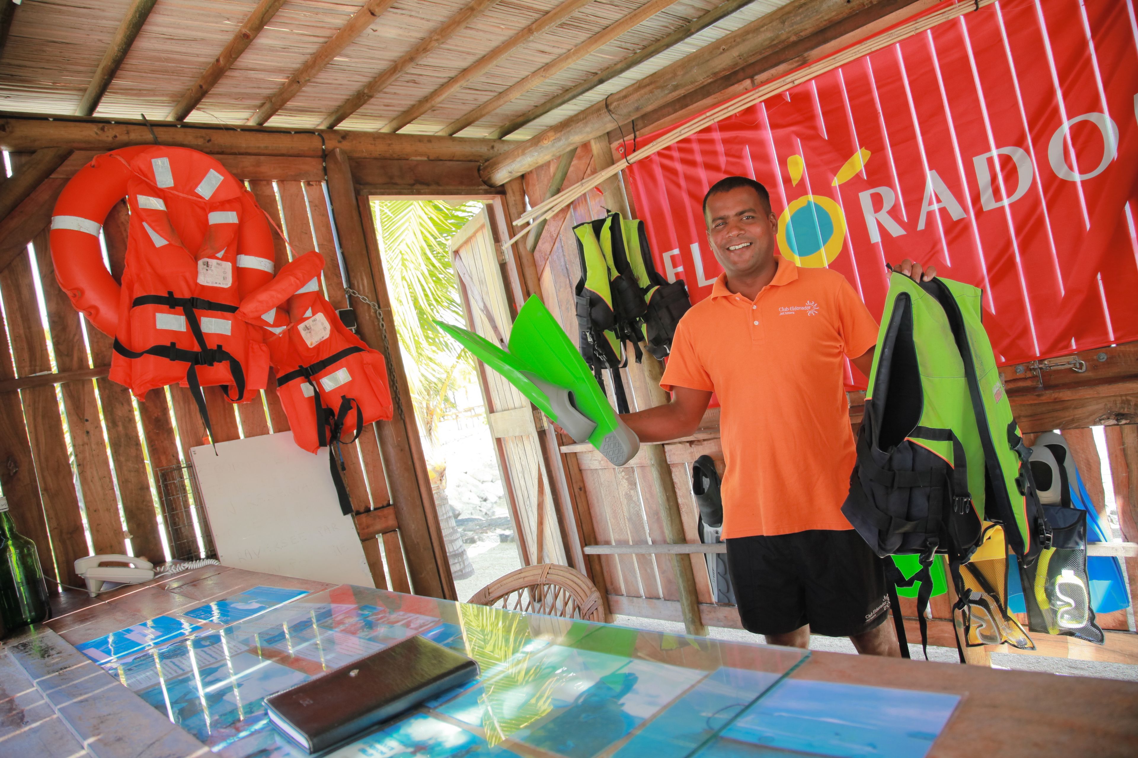 Un hombre con camisa naranja de pie junto a una mesa con chalecos salvavidas y una tabla de surf.