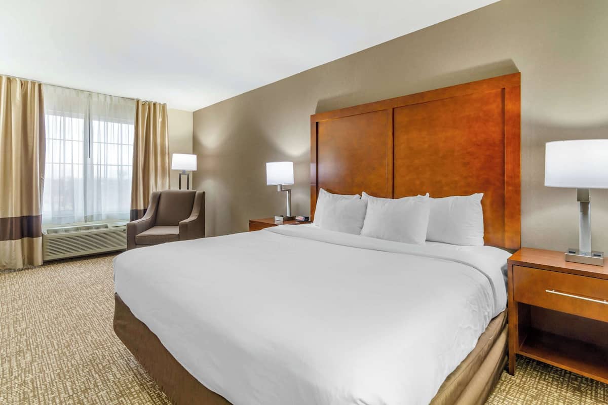 A hotel room with a large bed, two bedside tables with lamps, a chair, and a large window with curtains.