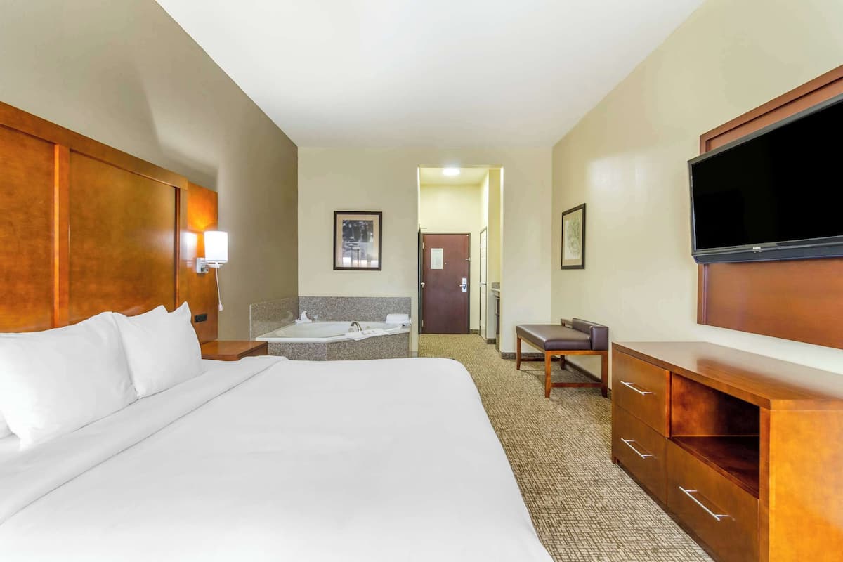 A hotel room with a large bed, a desk, a chair, a TV, a bathroom with a bathtub, and a door leading to another room.