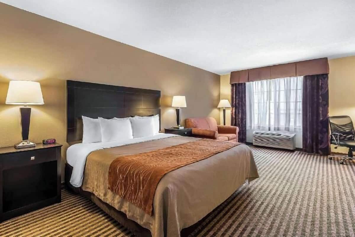 A hotel room with a large bed, two bedside tables with lamps, a chair, a window with curtains, and a carpeted floor.