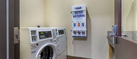 Un espace buanderie comprenant un lave-linge à chargement frontal, un distributeur de savon et d’eau de Javel fixé au mur, et un petit plan de travail.