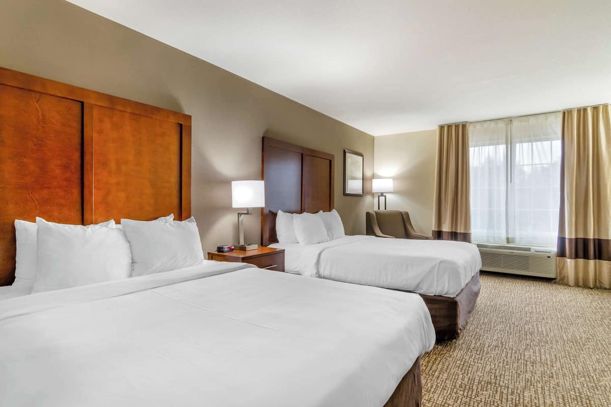 A hotel room with two beds, a wooden headboard, a desk with a lamp, a chair, and a large window with curtains.