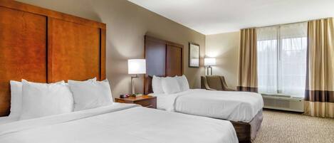 A hotel room with two beds, a wooden headboard, a desk with a lamp, a chair, and a large window with curtains.