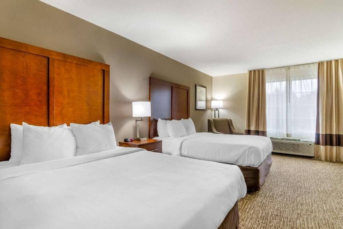A hotel room with two beds, a wooden headboard, a desk with a lamp, a chair, and a large window with curtains.