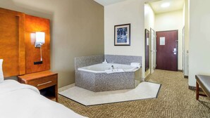 Quarto de hotel com uma banheira grande, mesa de cabeceira de madeira com abajur, quadro na parede e porta que dá para outro cômodo.