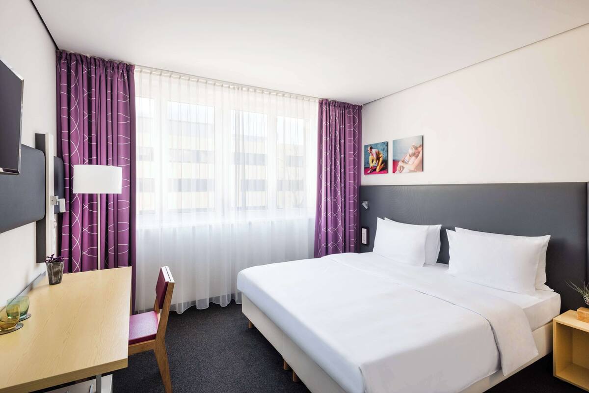Hotelkamer met een bed, bureau, stoel, televisie en raam met paarse gordijnen.