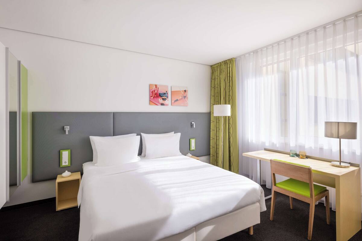 Een hotelkamer met een bed, bureau, stoel, lamp en een raam met gordijnen.