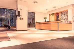 A hotel reception area with a wooden reception desk, a large window, and a bench with a bag on it. - Lindner Hotel Frankfurt Sportpark, part of JdV by Hyatt (Frankfurt)