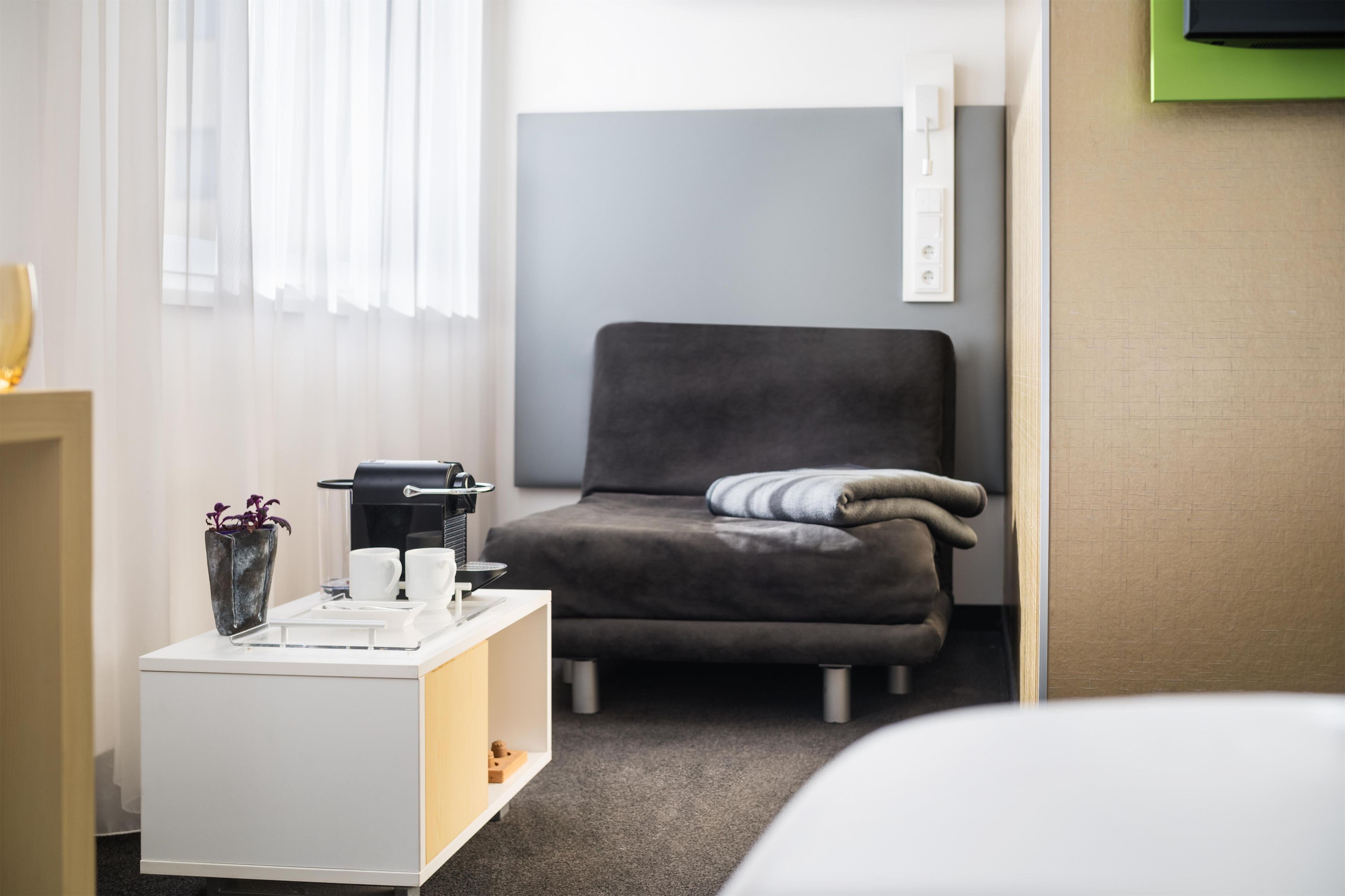 Ein modernes Hotelzimmer mit einem grauen Sofa, einem weißen Beistelltisch mit Kaffeemaschine und einer kleinen Pflanze.