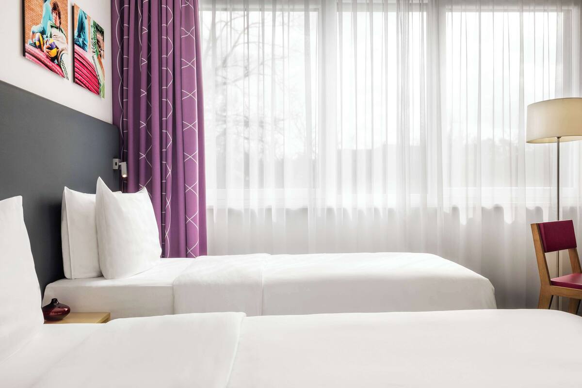 Een hotelkamer met twee bedden, een groot raam met dunne gordijnen, een vloerlamp en een stoel.
