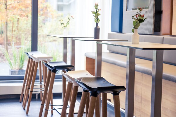 A modern dining area with a glass table, wooden chairs, and a vase with flowers. - Lindner Hotel Frankfurt Sportpark, part of JdV by Hyatt (Frankfurt)