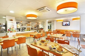 A modern cafeteria with orange chairs and tables, a kitchen area, and a television screen. - Suite Home Apt Luberon (Apt)