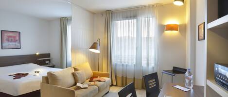 Quarto de hotel com cama, sofá, escrivaninha com cadeira, garrafa de água e televisão.