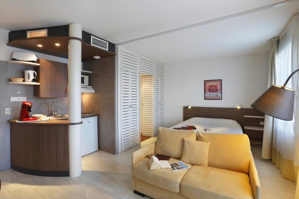 A compact hotel room with a kitchenette, a bed, a sofa, and a bedside lamp.