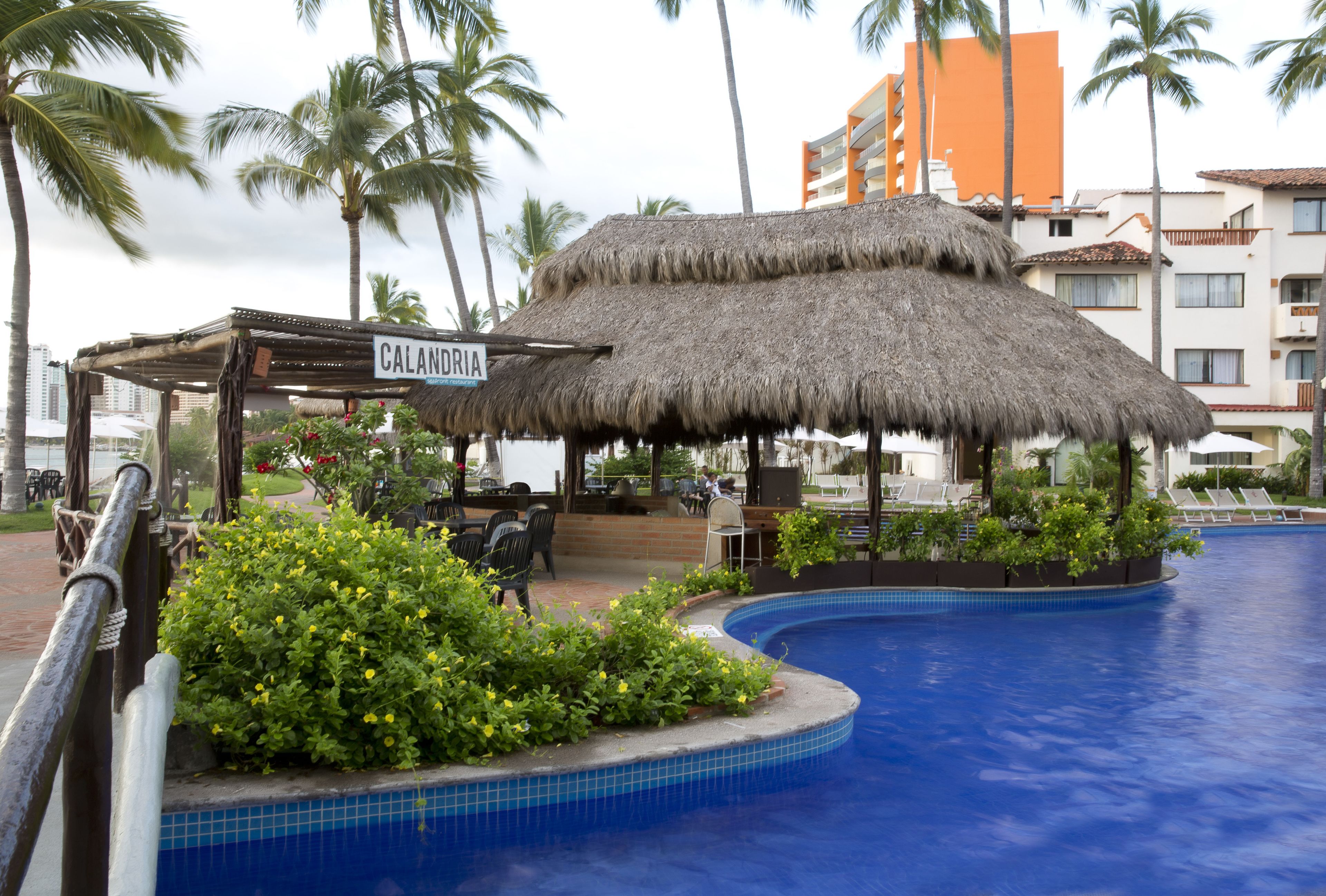 A pool area with a thatched-roof structure labeled "Calandria," surrounded by lush greenery and palm trees.