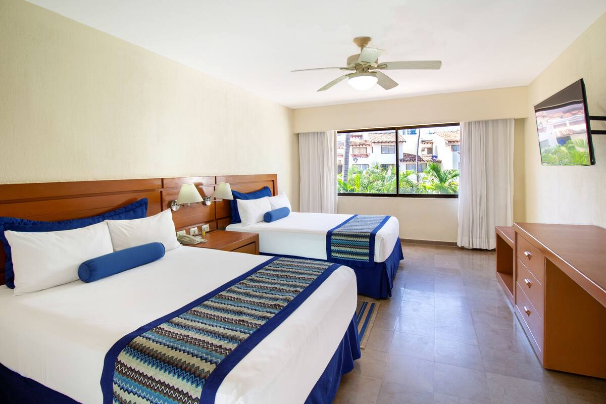 Habitación de hotel con dos camas, un escritorio, un ventilador de techo y una ventana con vistas a edificios y zonas verdes.