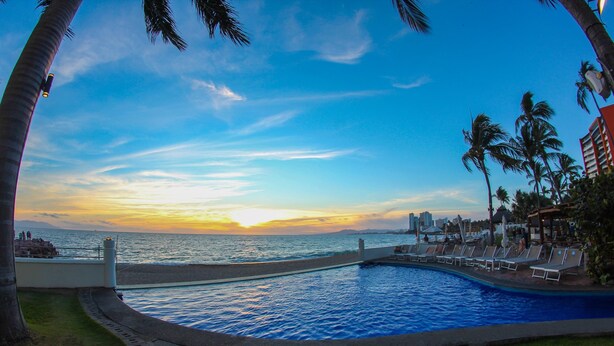 Una piscina con vistas a la puesta de sol sobre el océano, rodeada de palmeras y sillones de descanso.