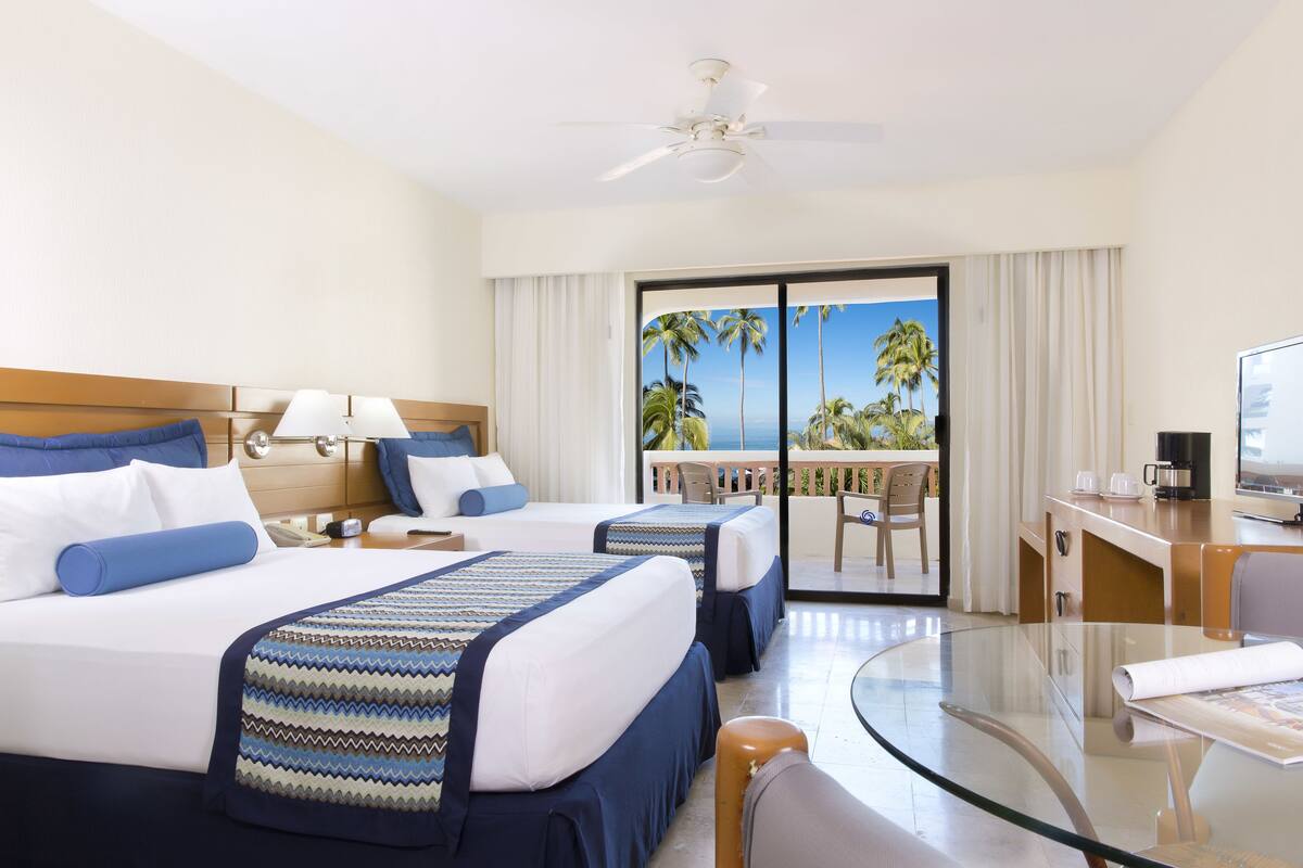 Habitación de hotel con dos camas, un escritorio, una silla, una mesita de vidrio y un balcón con vista a palmeras y al océano.