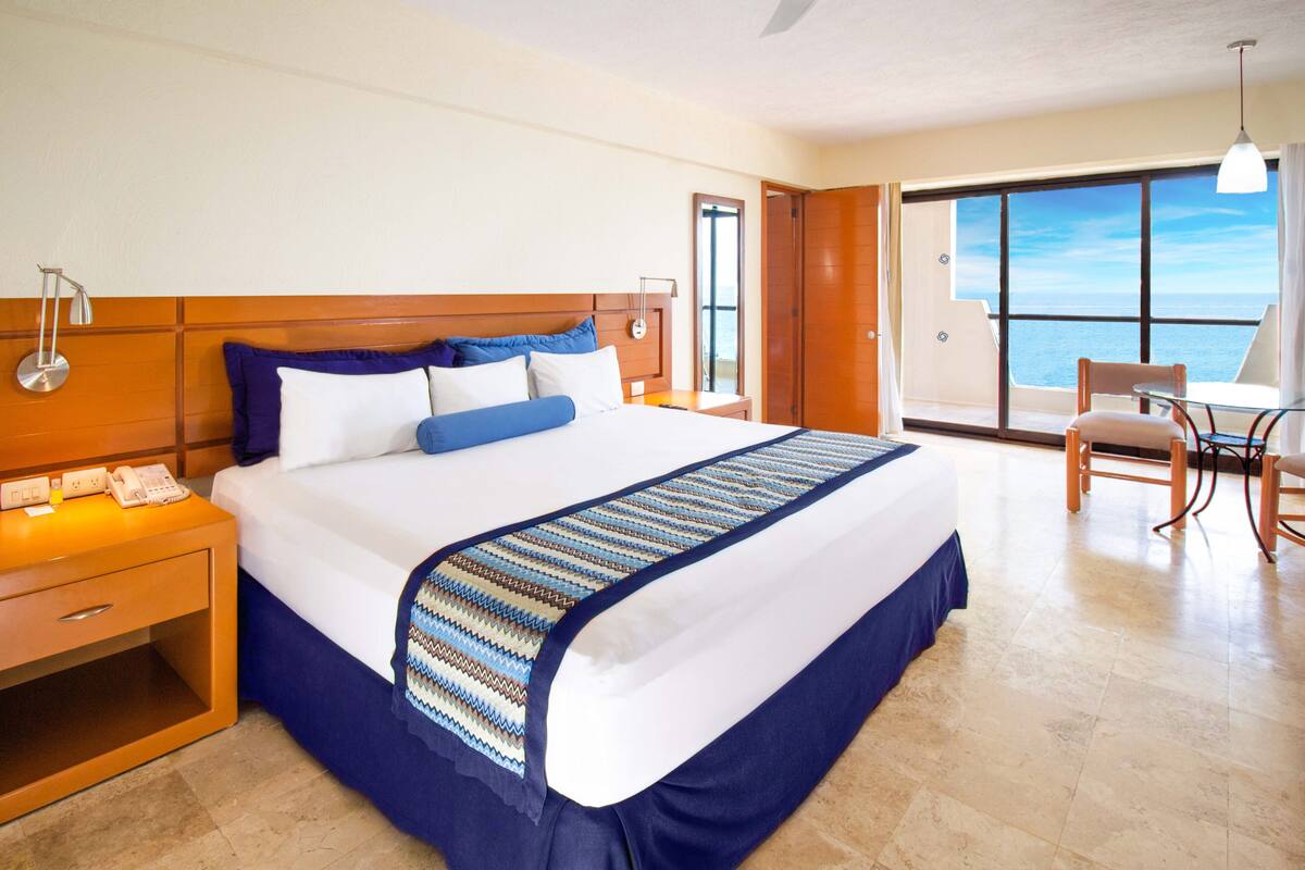 Una habitación de hotel con una cama grande, una mesita de noche con teléfono, una silla y vistas al mar a través de un ventanal amplio.