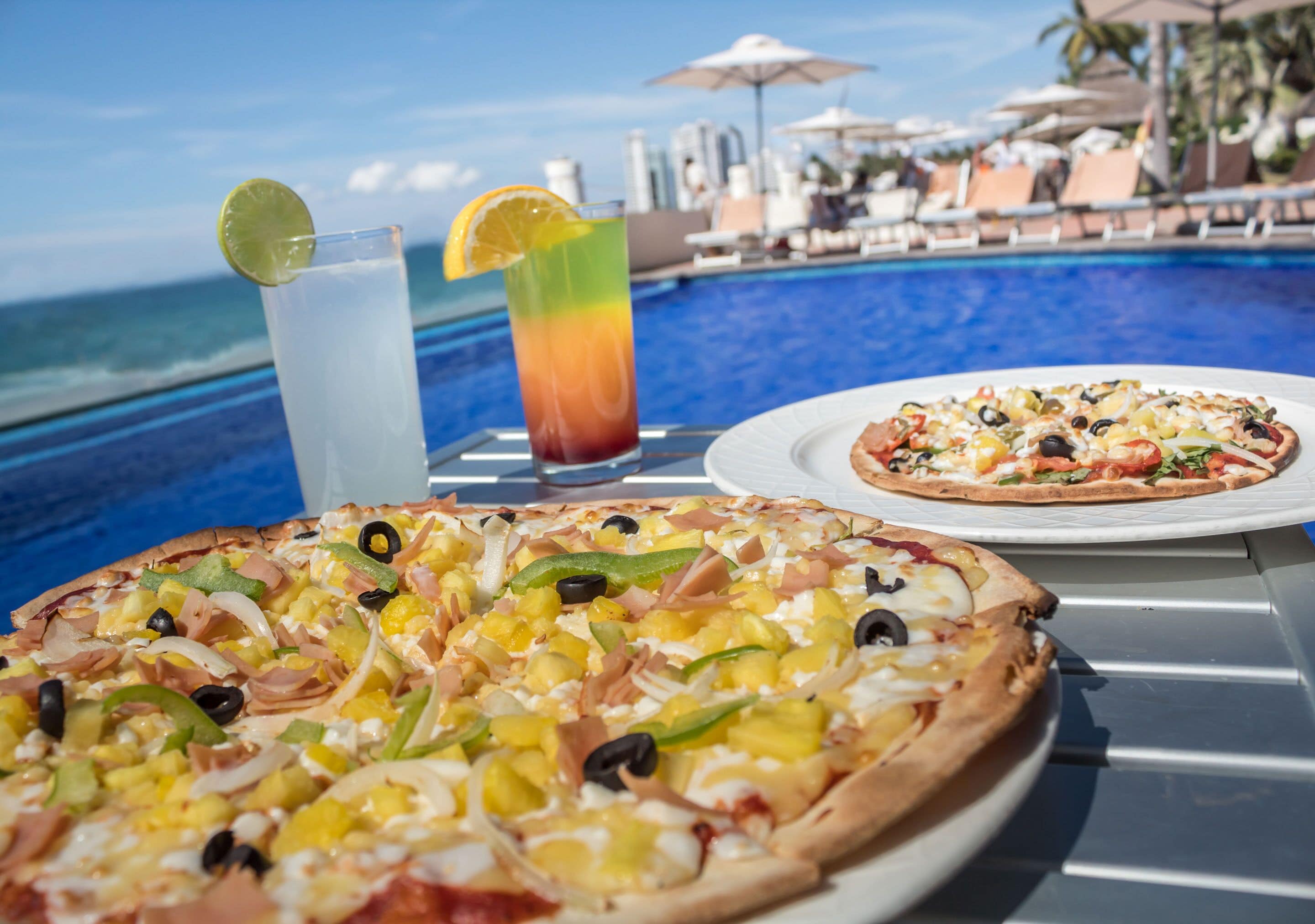 Mesa com duas pizzas e duas bebidas à beira da piscina.